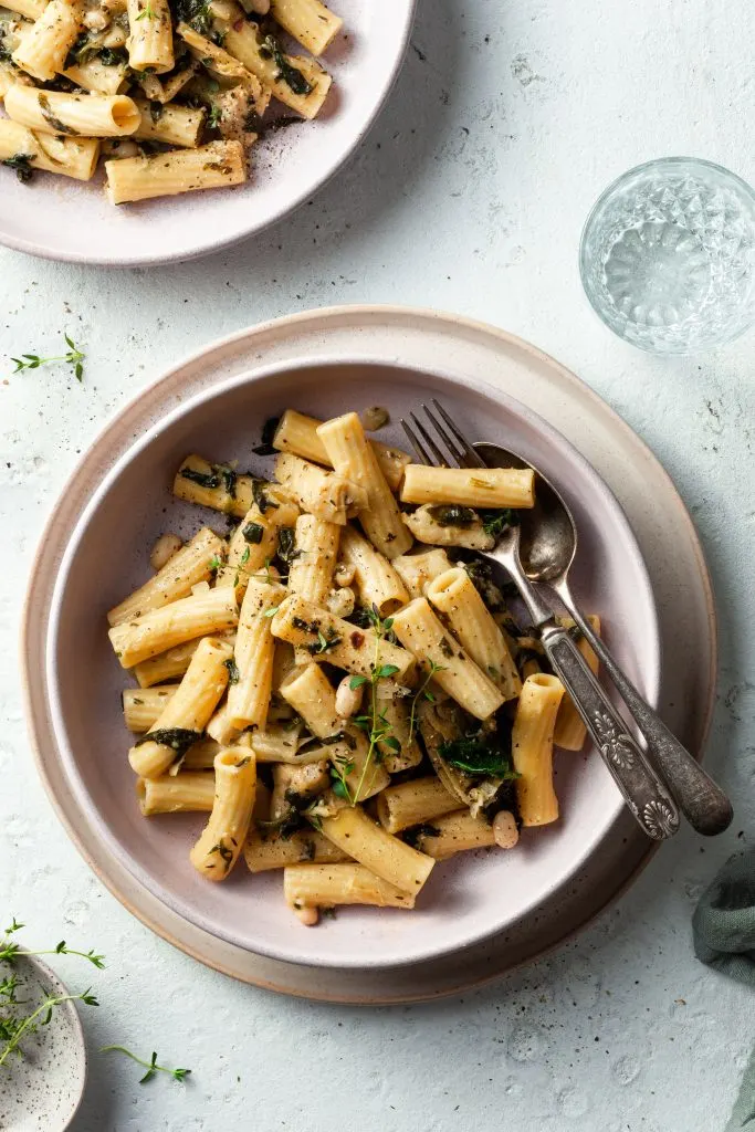A pink bowl filled with gemelli noodles, spinach, artichokes, fresh thyme and white beans.