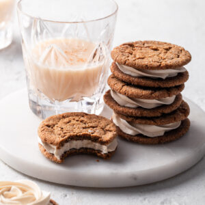A stack of molasses sandwich cookies beside a glass of vegan eggnog all on a white marble plate