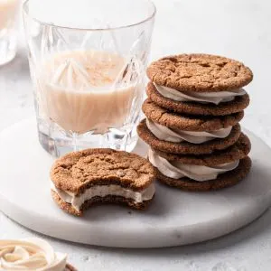 A stack of molasses sandwich cookies beside a glass of vegan eggnog all on a white marble plate