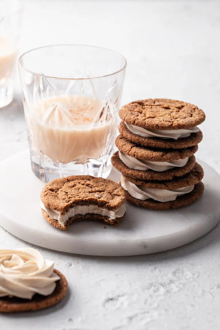 A stack of molasses sandwich cookies beside a glass of vegan eggnog all on a white marble plate