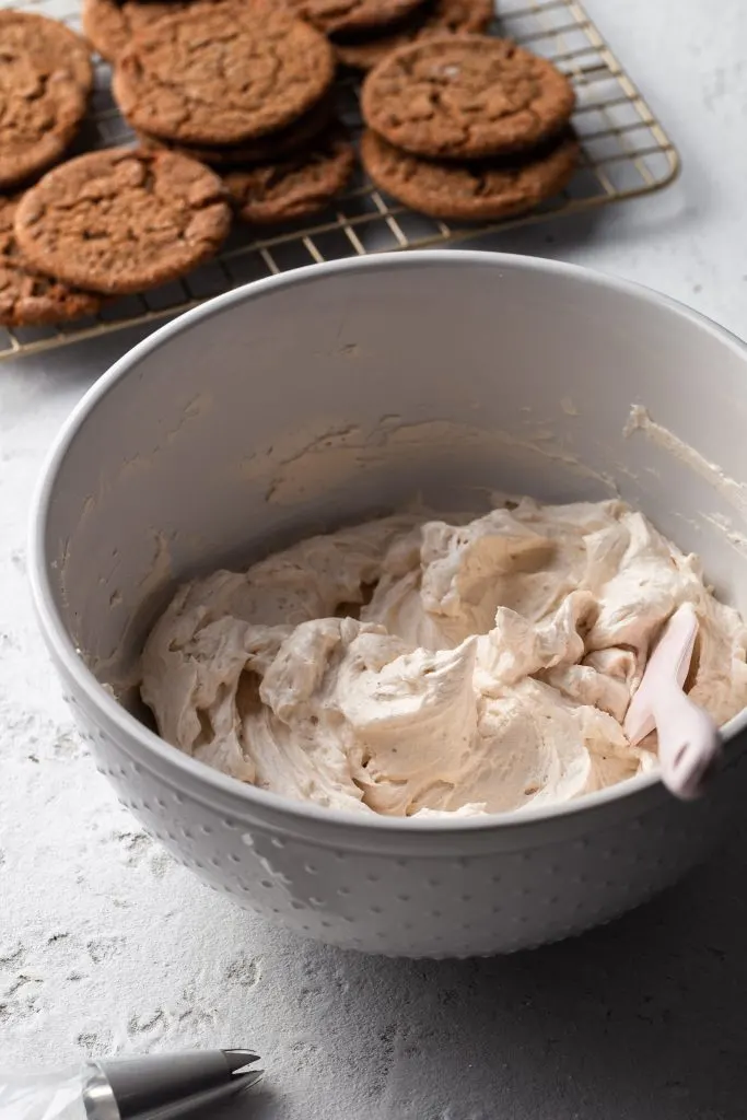 Eggnog frosting in a white bowl beside a piping bag. In the background is a cooling rack of molasses cookies