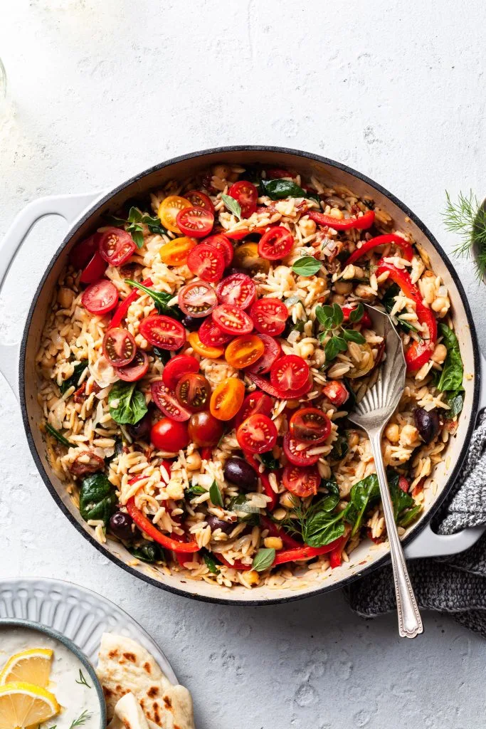A white enamel skillet filled with orzo, olives, sliced red peppers, and veggies. 