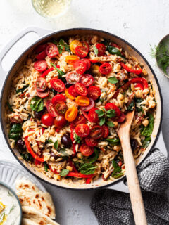 A white cast iron skillet filled with orzo, chickpeas, tomatoes and other Greek inspired herbs and veggies.