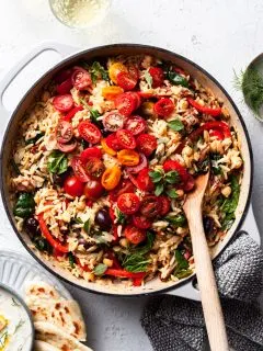 A white cast iron skillet filled with orzo, chickpeas, tomatoes and other Greek inspired herbs and veggies.
