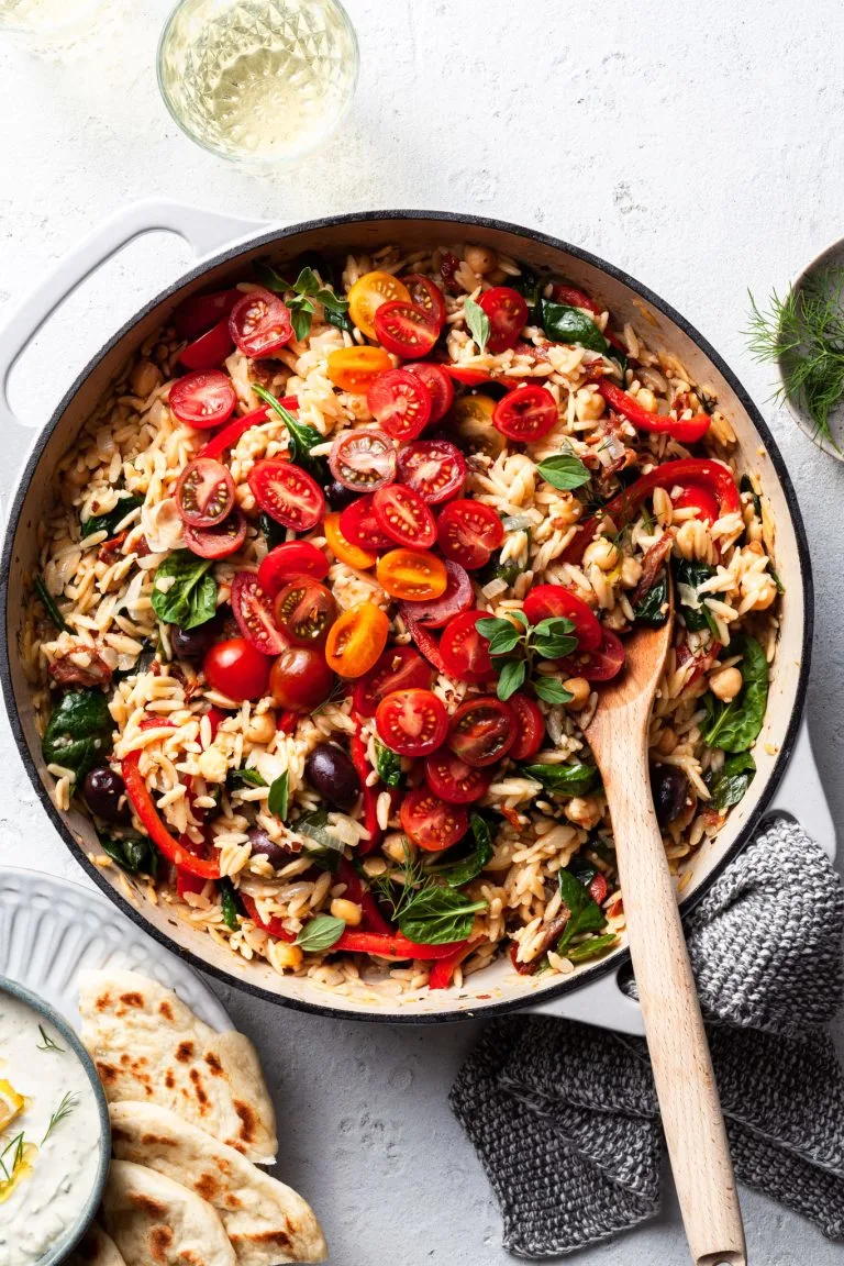 A white cast iron skillet filled with orzo, chickpeas, tomatoes and other Greek inspired herbs and veggies.