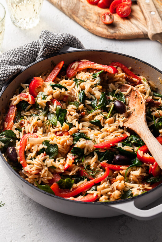 A white skillet filled with orzo, red capsicums, olives, spinach and dill. Sliced cherry tomatoes lay on a cutting board in the background. 