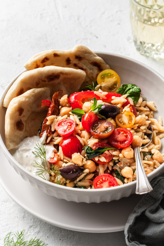 A white bowl filled with orzo, olives, chickpeas, herbs, cherry tomatoes, a side of vegan tzatziki, and flatbread. 