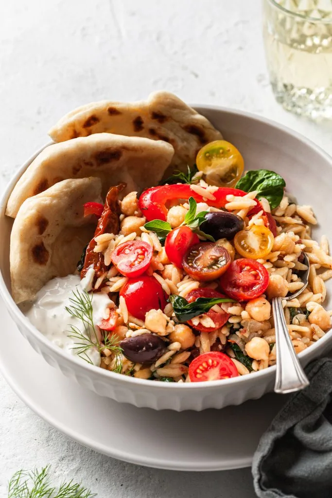 A white bowl filled with orzo, olives, chickpeas, herbs, cherry tomatoes, a side of vegan tzatziki, and flatbread. 