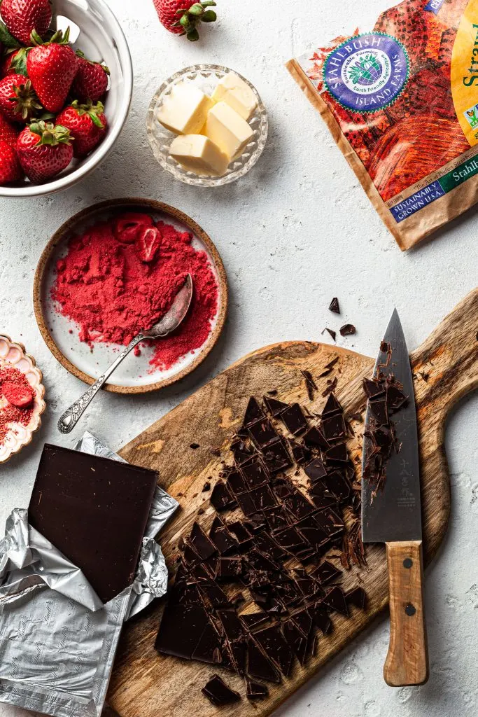 W hite counter covered with the measured ingredients for strawberry truffles, including chopped chocolate on a cutting board, a plate of crushed strawberries, and basket of strawberries and a small bowl of butter. 