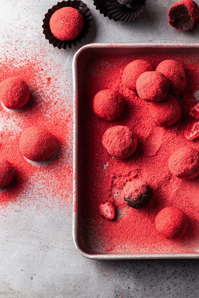 An aluminum tray filled with crushed bright pink crushed freeze dried strawberries, and chocolate truffles being rolled in the strawberry coating. 