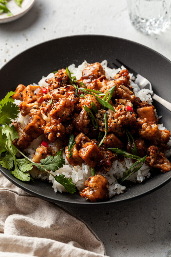 Side view of a black bowl filled with cubes of tofu and cauliflower florets in a stick dark sauce, served over rice. 