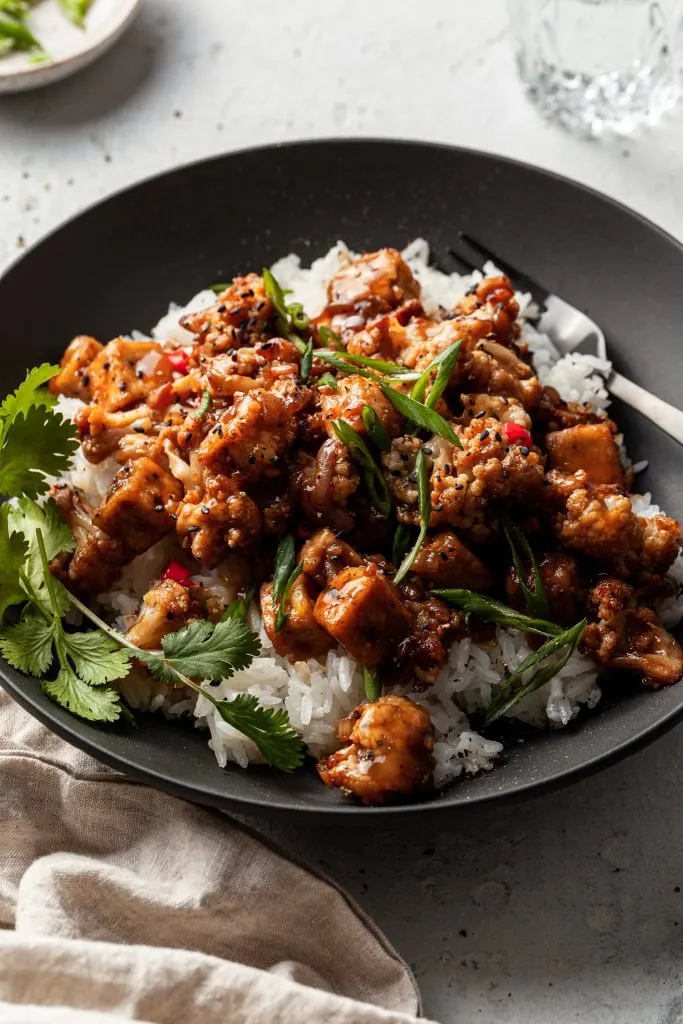 Side view of a black bowl filled with cubes of tofu and cauliflower florets in a stick dark sauce, served over rice. 