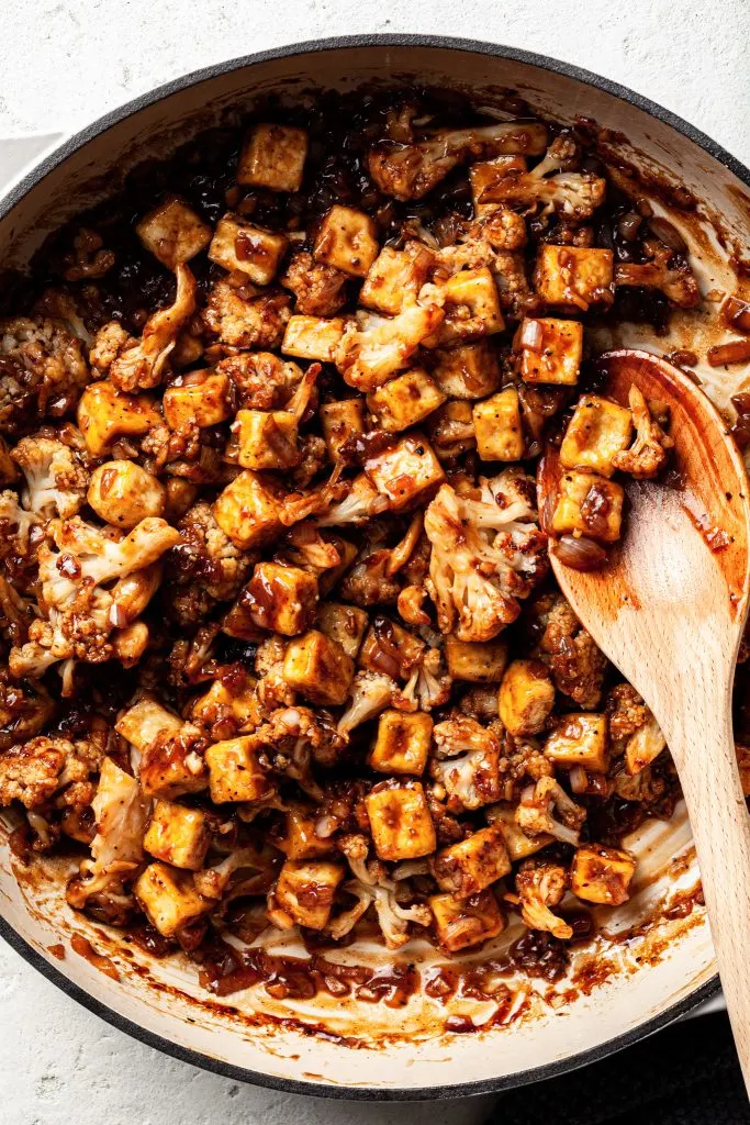 White enamel skillet filled with crispy tofu cubes and roasted cauliflower all coated in a sticky dark sauce. 