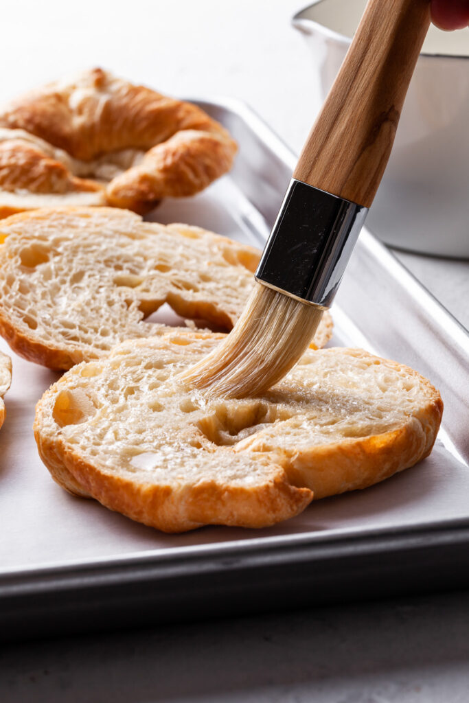 A pastry brush is brushing simple syrup onto the sliced surface of a croissant.  