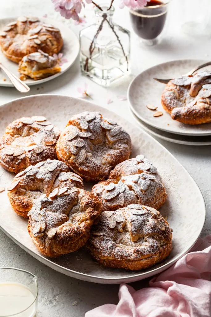 A cream platter filled with almond croissants, beside a stack of cream plates, a vase of spring cherry blossoms and a cup of coffee.