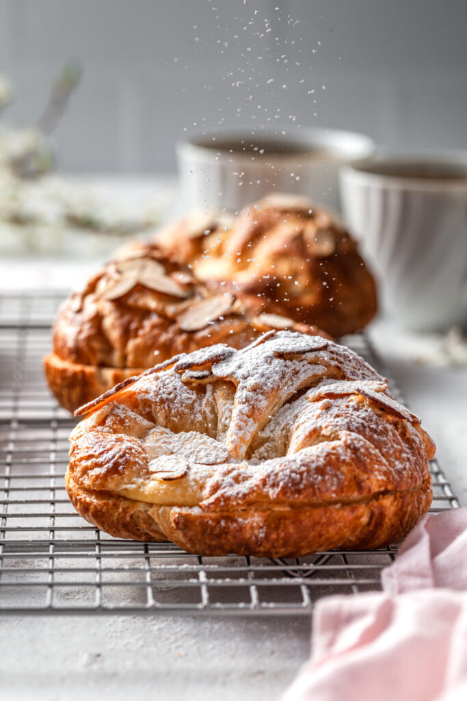 An almond croissant on a cooling rack being dusted with powdered sugar. 