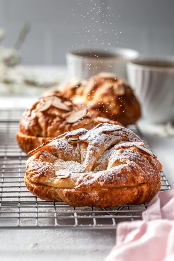 An almond croissant on a cooling rack being dusted with powdered sugar. 
