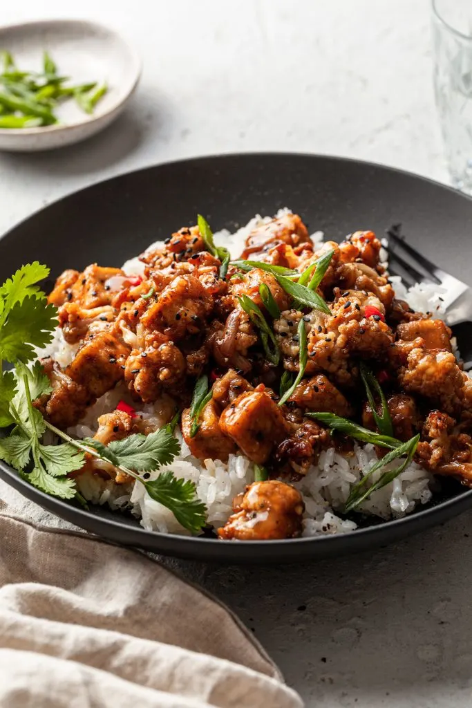 A black bowl filled with white rice topped with crispy tofu, cauliflower and a black pepper sauce. Garnished with sliced green onions. 