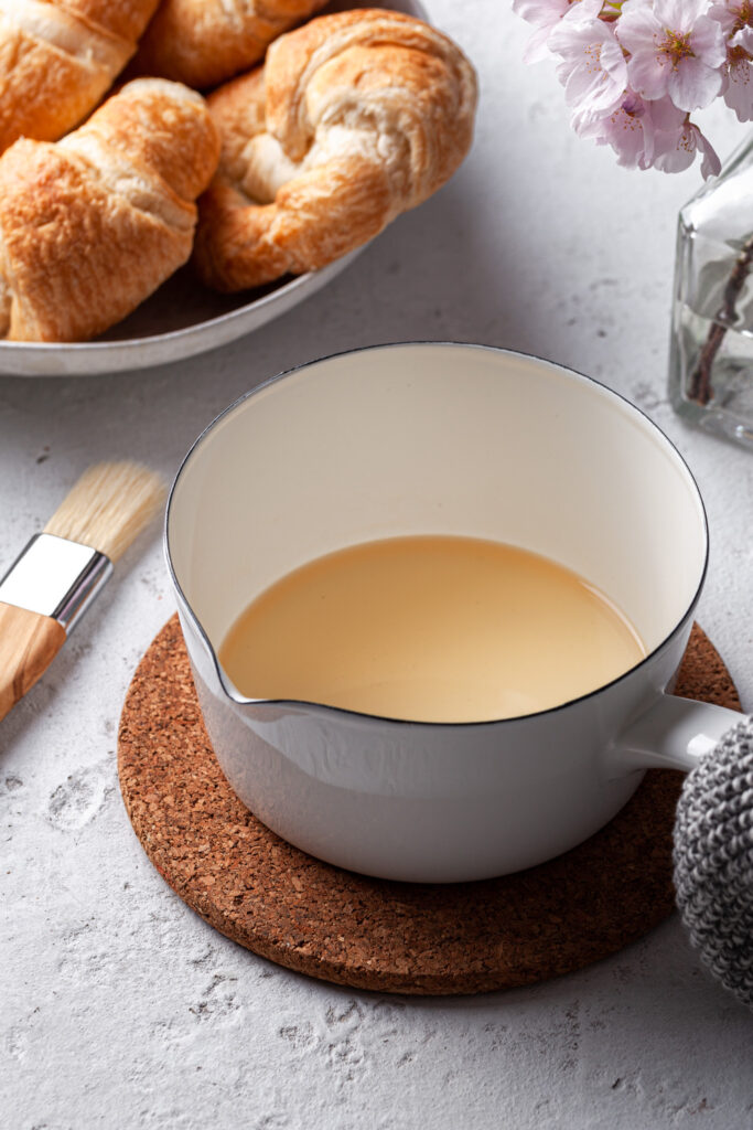A small white pot with simple syrup in it. In the background there's a cream platter filled with croissants, beside a pastry brush. 