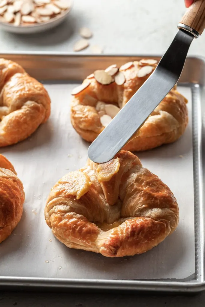 An offset spatula spreads a small amount of frangipane to the top of the almond croissant. 