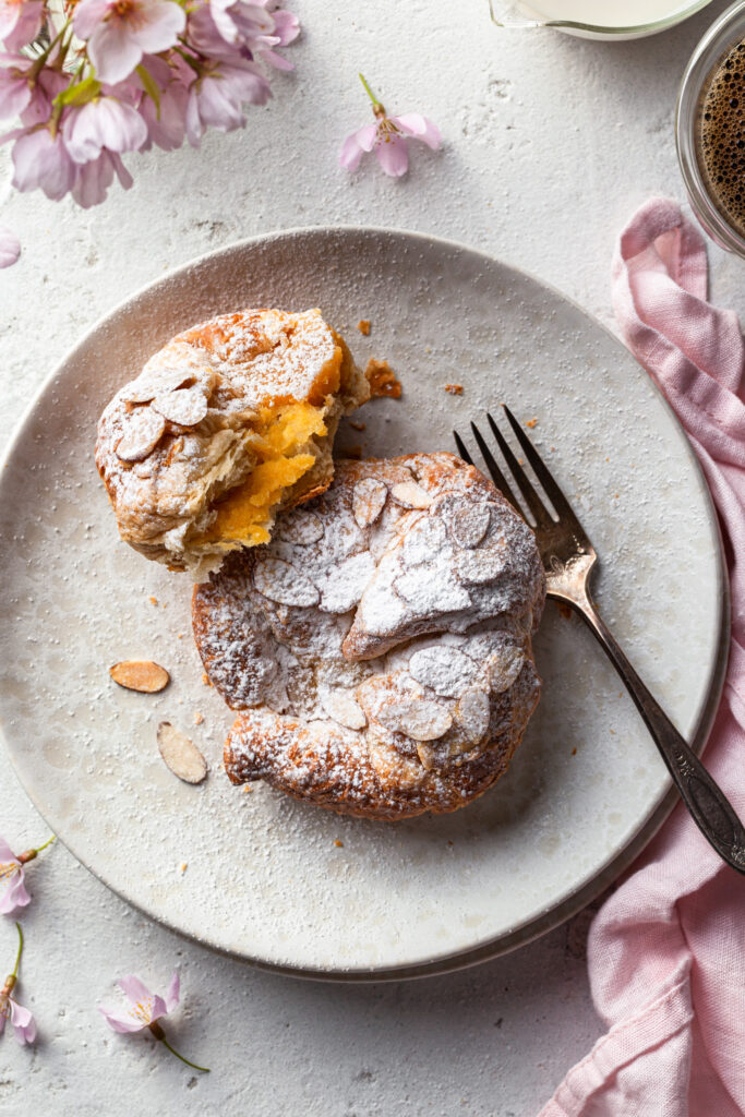 A cream plate filled with a whole vegan almond croissant and a partially eaten one revealing the creamy almond filling. 