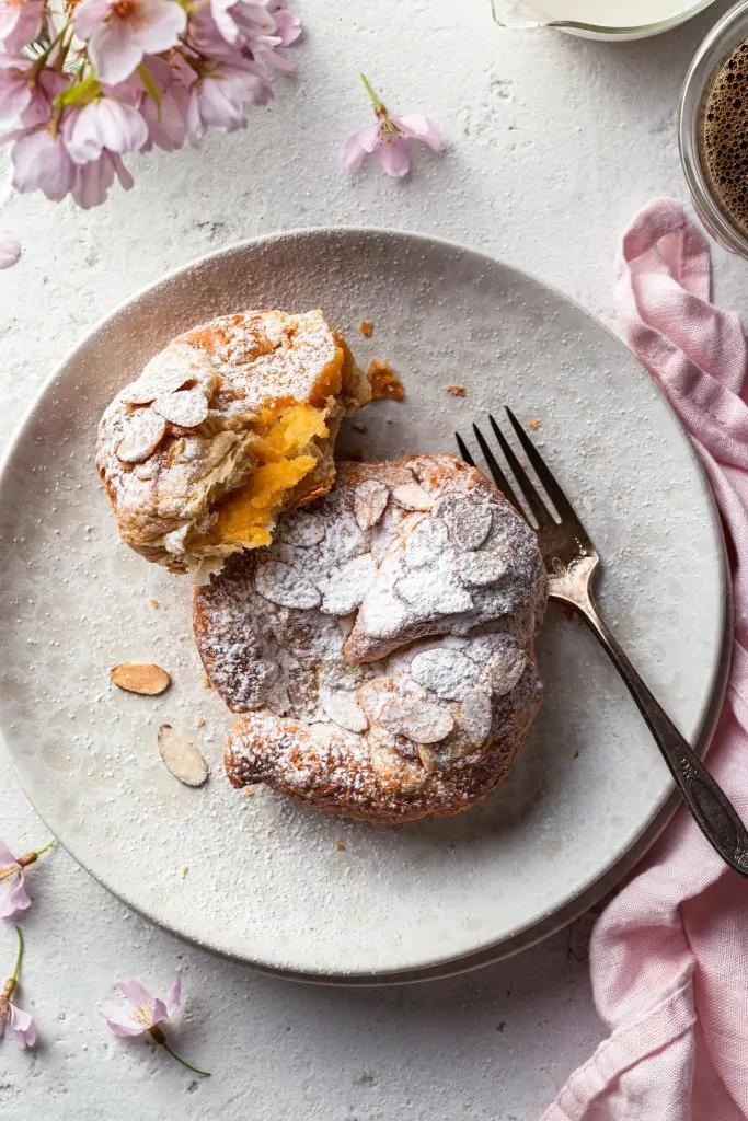 A cream plate filled with a whole vegan almond croissant and a partially eaten one revealing the creamy almond filling. 