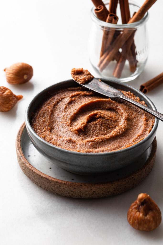 A blue bowl containing fig filling for sticky buns. Dried figs are scattered around the bowl, and cinnamon sticks are in a glass in the background. 