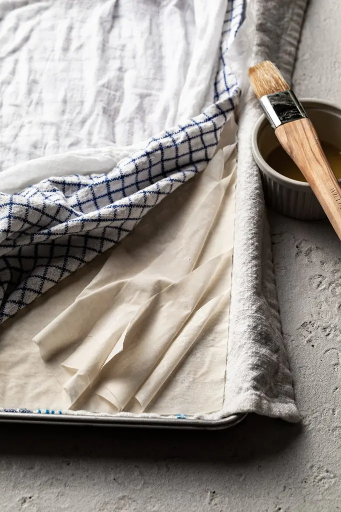 Sheets of phyllo dough on a towel lined baking tray, covered with a second kitchen towel, which is pulled back a bit to expose the layers of pastry. 