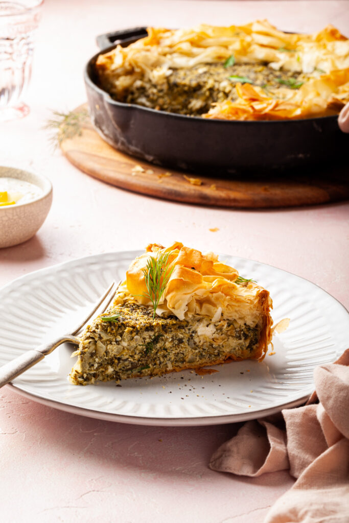 A piece of spanakopita served on a white plate in front of a cast-iron skillet where the rest of the spanakopita is. 