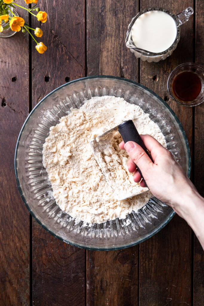 A glass bowl filled with a crumbly flour mixture and pea sized pieces of vegan butter being cut in with a pastry cutter. 