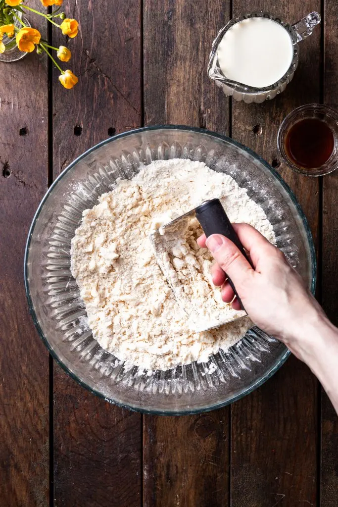 A glass bowl filled with a crumbly flour mixture and pea sized pieces of vegan butter being cut in with a pastry cutter. 