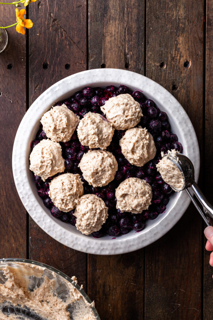 Drop biscuit batter being scooped over a frozen blueberry filling in a white round baking dish. 