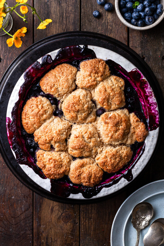 Blueberry cobbler fresh out of the oven in a white round baking dish, on a dark wooden table. 