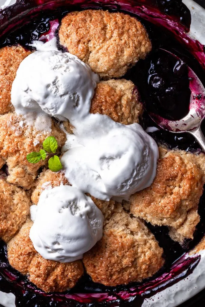 Close up of a blueberry cobble in a white baking dish with 3 scoops of melting vanilla ice cream. 
