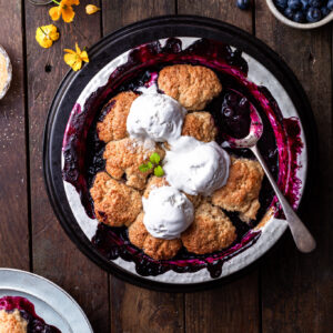 Baked blueberry cobbler in a white round baking dish, with 3 scoops of vanilla ice cream served on top.