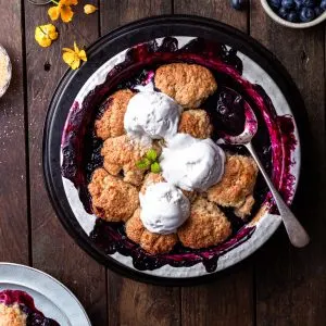 Baked blueberry cobbler in a white round baking dish, with 3 scoops of vanilla ice cream served on top.
