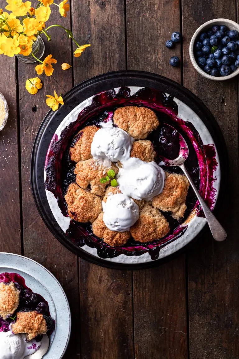 Baked blueberry cobbler in a white round baking dish, with 3 scoops of vanilla ice cream served on top.