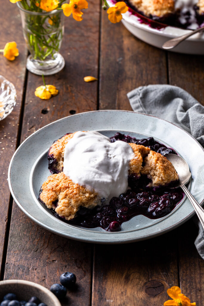 A serving of berry cobbler on a light blue plate topped with melting whip, and one of the biscuits is cup open to reveal the tender interior. 