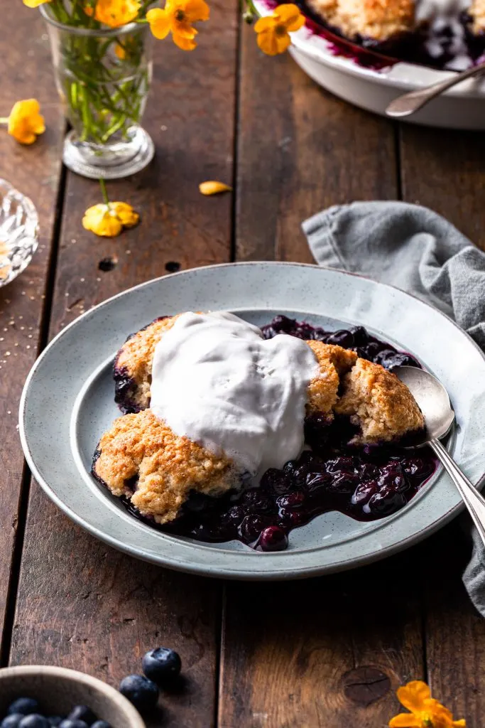 A serving of berry cobbler on a light blue plate topped with melting whip, and one of the biscuits is cup open to reveal the tender interior. 