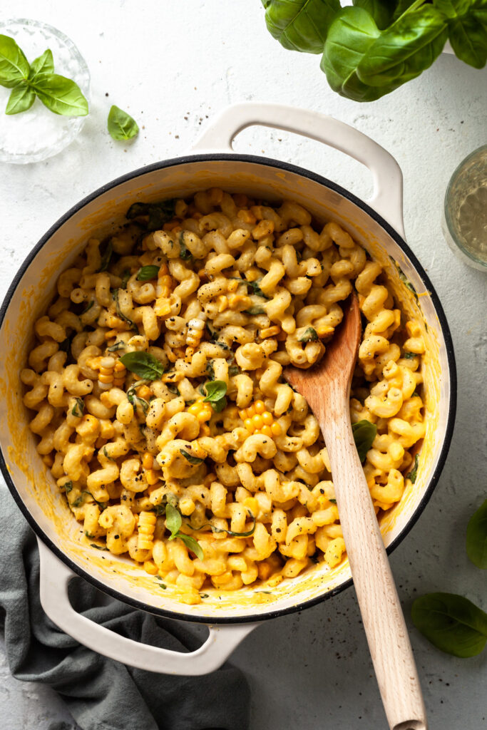 A white enamel pot filled with curly pasta noodles covered in a yellow sauce, with corn and herbs. 