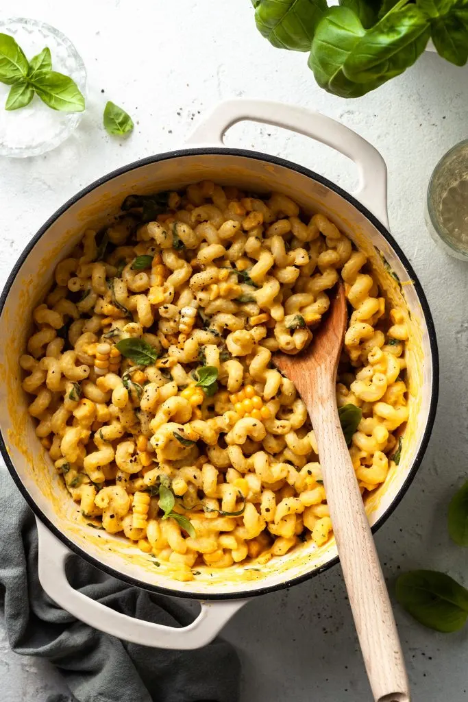A white enamel pot filled with curly pasta noodles covered in a yellow sauce, with corn and herbs. 