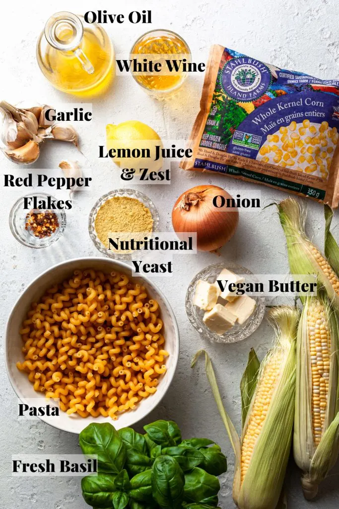 A white counter covered in ingredients measured out in glass and white ceramic bowls, such as pasta, corn, nutritional yeast, garlic, etc. 