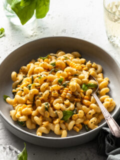 A light grey coup plate filled with cavatappi pasta coated in a creamy sauce studded with corn kernels and basil.