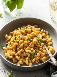 A light grey coup plate filled with cavatappi pasta coated in a creamy sauce studded with corn kernels and basil.