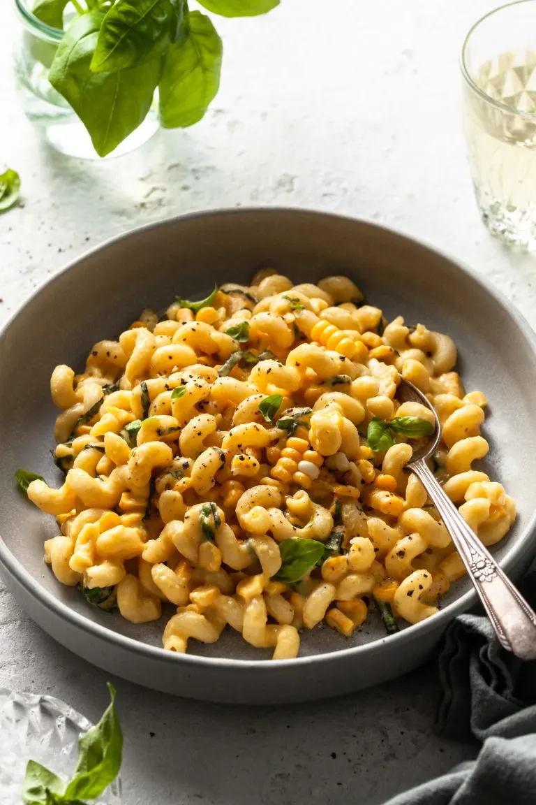 A light grey coup plate filled with cavatappi pasta coated in a creamy sauce studded with corn kernels and basil.