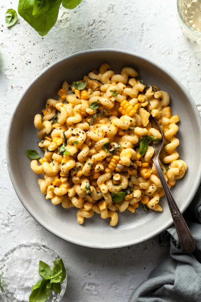 A light grey bowl filled with corkscrew pasta coated in a light yellow cream sauce, mixed with corn kernels and basil leaves. 