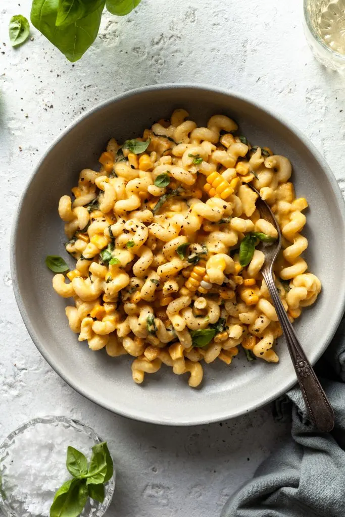 A light grey bowl filled with corkscrew pasta coated in a light yellow cream sauce, mixed with corn kernels and basil leaves. 