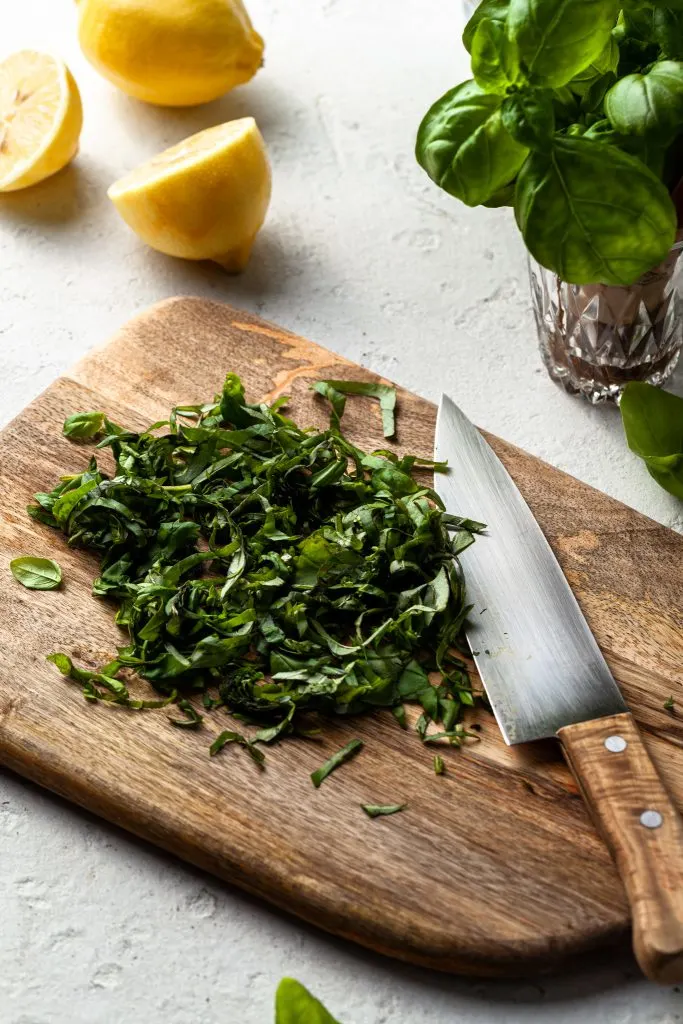 A pile of julienned basil leaves on a wooden cutting board, and lemons in the background. 