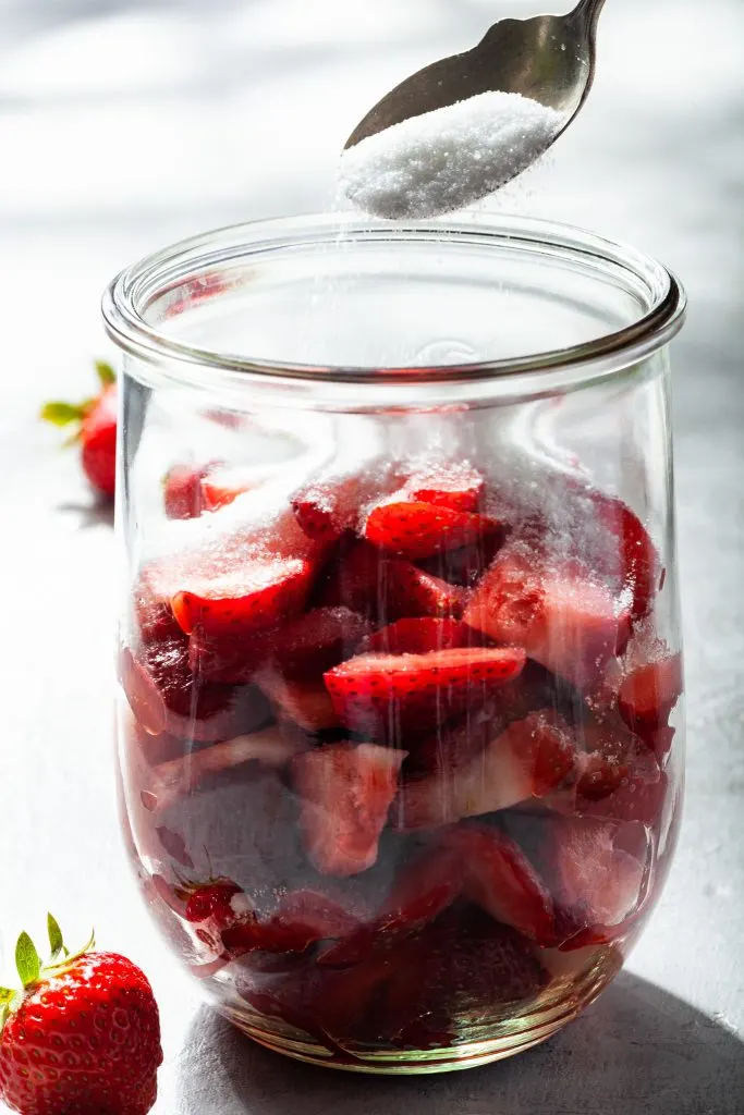A large glass jar filled with sliced strawberries, and sugar being spooned in over the berries.