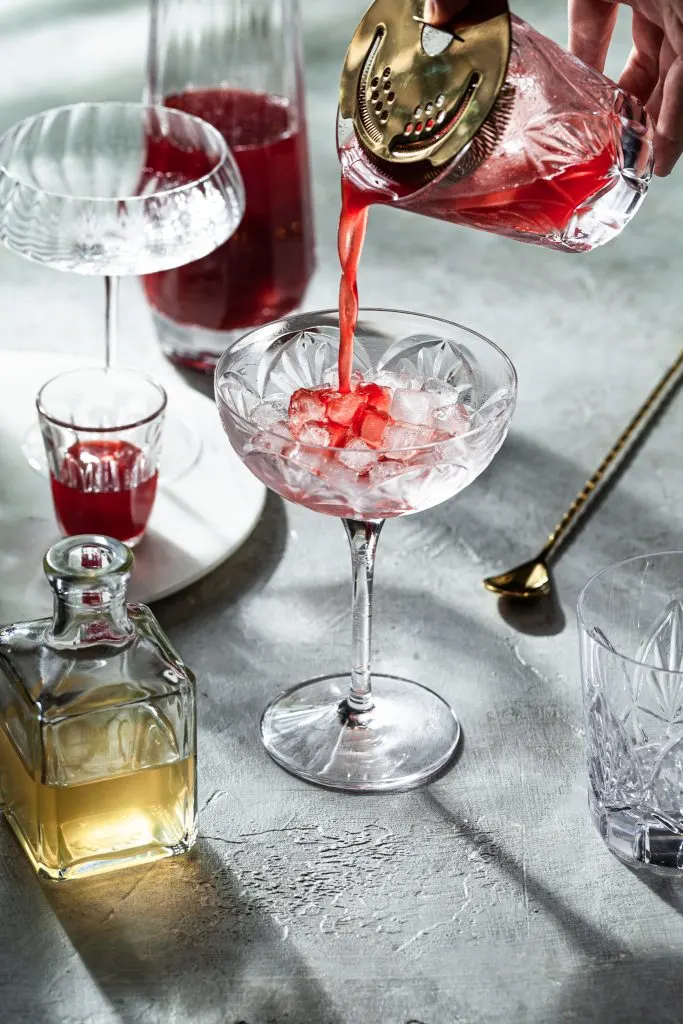 A glass with brass Hawthorne strainer straining a red drink (shrub syrup) into a stemmed coup glass filled with ice.